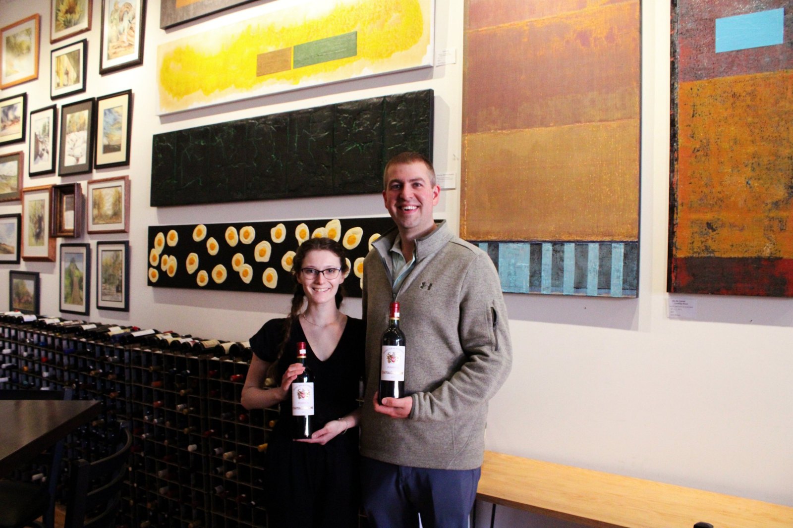 Two people holding bottles of wine in front of original art at 2 Roots Gallery
